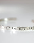 A silver cuff bracelet resting on a white surface, showing the stamped text METTA and May all.