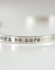 A silver metal cuff bracelet angled to display the hand-stamped phrase be safe.