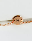 A rose gold chain bracelet featuring a small circular disk engraved with the letter H.