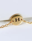 A close-up view of a gold circular charm engraved with the letter H on a delicate gold chain.