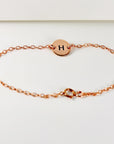A full view of a rose gold link bracelet with a centered circular disk charm engraved with an H and a spring-ring clasp.