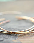 Three thin metal open cuff bracelets in silver, rose gold, and yellow gold are stacked on a rough gray surface.