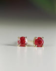 Two round red ruby stud earrings in four-prong gold settings standing upright on a light surface.
