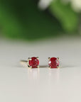 Two round red ruby stud earrings displayed side-by-side on a light surface against a blurred green background.