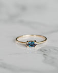 A gold ring with a horizontally set marquise-cut blue and purple gemstone resting on a white marble surface.