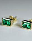 A pair of rectangular green stone stud earrings with gold bezel settings resting on a white surface at an angle.