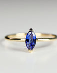 A thin gold ring featuring a prong-set marquise-cut blue sapphire solitaire, shown from the front on a light surface.