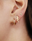 Side view of an earlobe showing a gold diamond huggie hoop earring alongside a lightning bolt stud and a bezel-set round stone stud.
