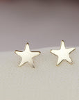 Two gold star stud earrings rest side by side on a soft white fabric surface.