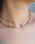 Detailed view of a person wearing a layered rose gold necklace set with a chunky paperclip chain and finer chain strands with a disc pendant.