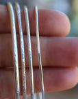 Close-up of four thin silver hammered cuff bracelets held vertically across a person fingers.