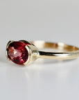 A side view of a gold ring with an east-west set oval pink tourmaline gemstone in a half-bezel setting against a plain light background.