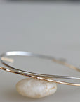 Two thin metal open cuff bracelets balance on top of a smooth light-colored stone against a plain light background.