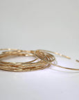A large stack of thin yellow gold hammered cuff bracelets is arranged on a smooth white surface.