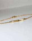 An angled shot of a thin gold chain bracelet showcasing a customized bar pendant with engraved text.