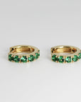 Close-up of two gold huggie hoop earrings lined with bright green stones, angled slightly toward each other.