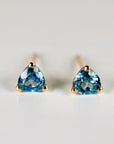 Front view of two trillion-cut blue gemstone stud earrings with gold prongs and posts on a white surface.