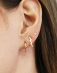 Close-up of an ear with three piercings wearing a small diamond huggie hoop, a gold lightning bolt stud, and a tiny stone stud.
