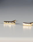 Two gold bar stud earrings are positioned on a grey surface, viewed from a low angle.