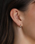 A close-up of a person wearing a small gold hoop earring with a bezel-set single solitaire diamond.