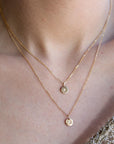 A person wearing a layered gold necklace set consisting of a short chain with an initial pendant and a longer chain with a bee pendant.