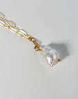 Angled close-up of a gold paperclip chain and white baroque pearl pendant resting on a light gray surface.