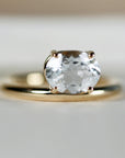 A gold ring with an east-west set, faceted clear oval gemstone rests on a light background.