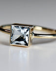Close up angled view of a gold ring featuring a bezel set square light blue gemstone on a white surface.