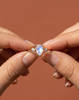 Two hands hold up the gold pear moonstone and diamond ring against a terracotta-colored background.