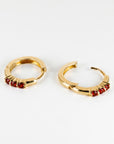 Two gold huggie hoop earrings shown close-up on a white surface, angled to display their three red garnet stones.