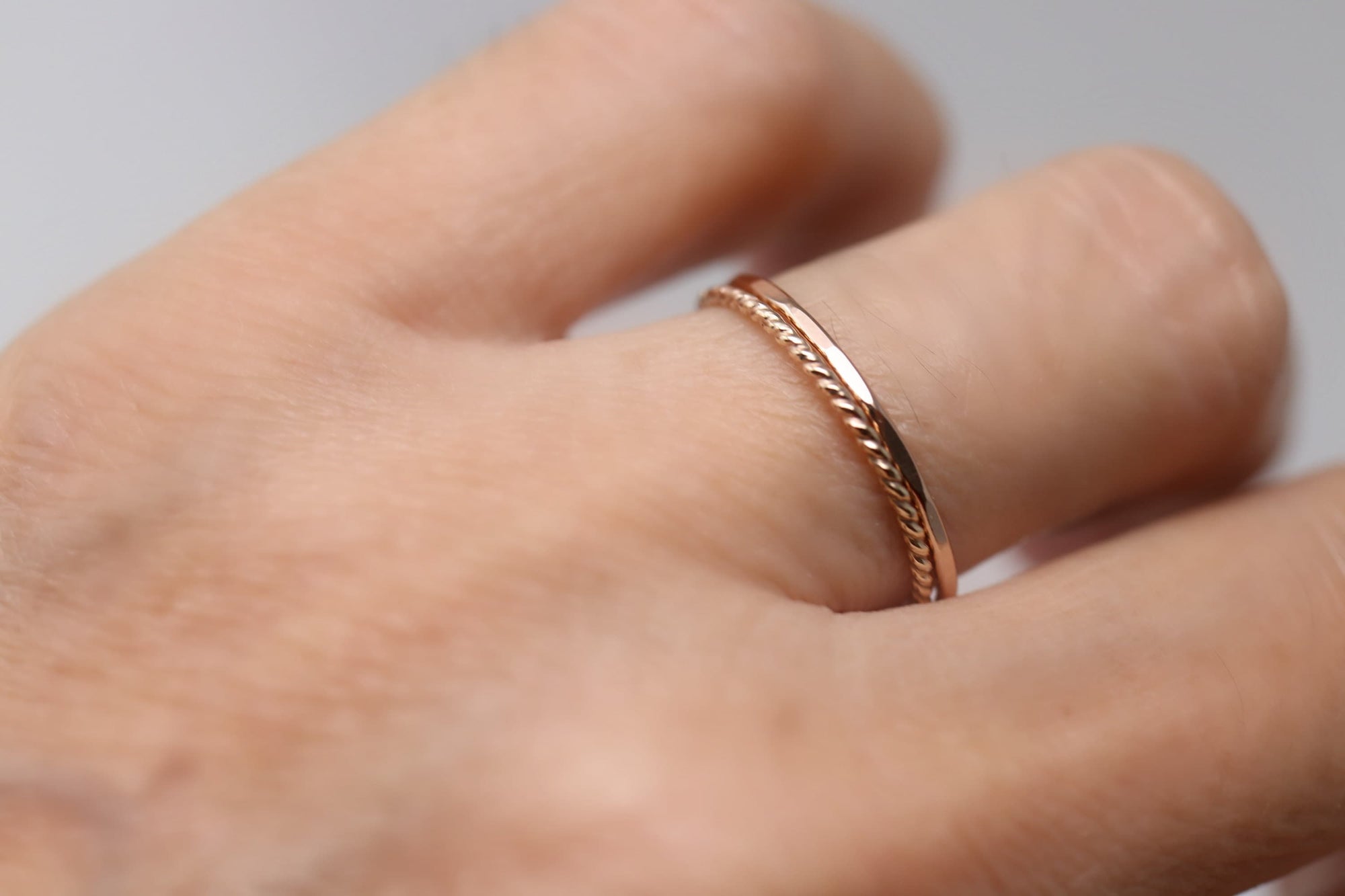 Two rose gold rings are shown on a finger. One ring is a simple band, and the other has a twisted rope design. The rings are delicate and appear to be made of a shiny metal.
