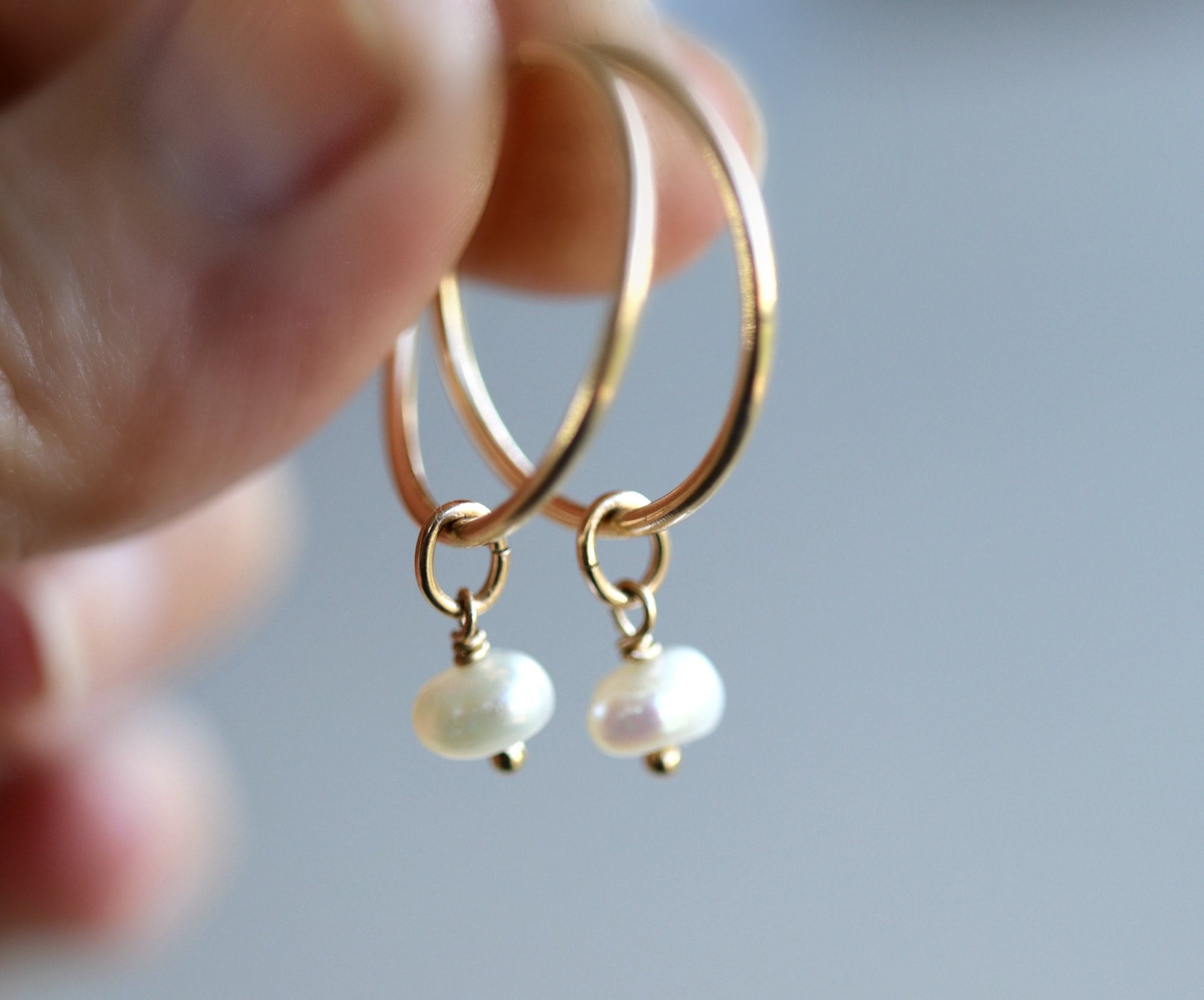 A pair of gold hoop earrings with small, white pearl drops. The earrings are held up against a soft, blurred background. The hoops are thin and shiny, and the pearls have a subtle luster.