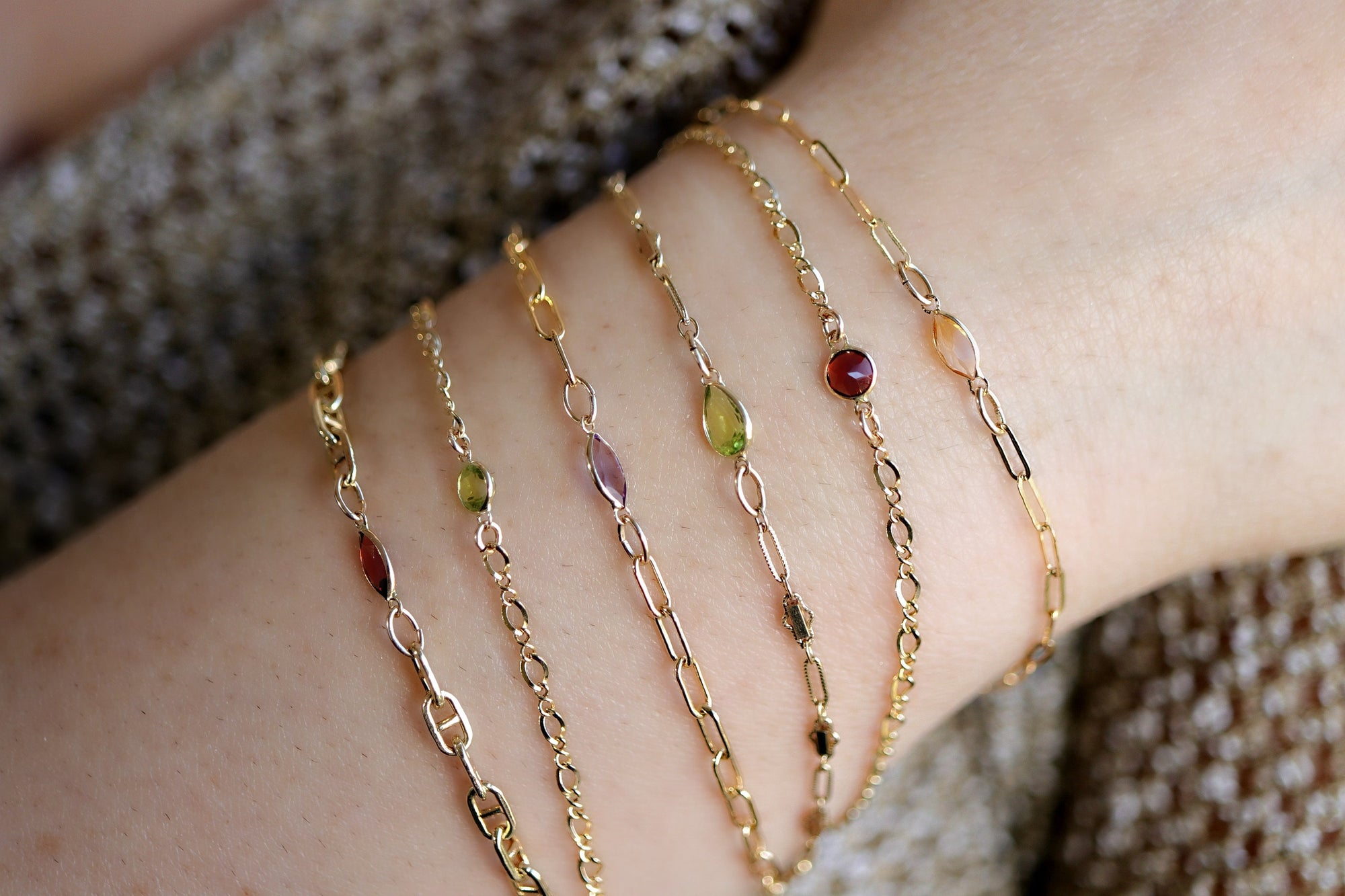 Five gold chain bracelets with different colored gemstones. The bracelets feature a delicate chain design and small, oval-shaped gemstones in shades of red, green, purple, and orange.