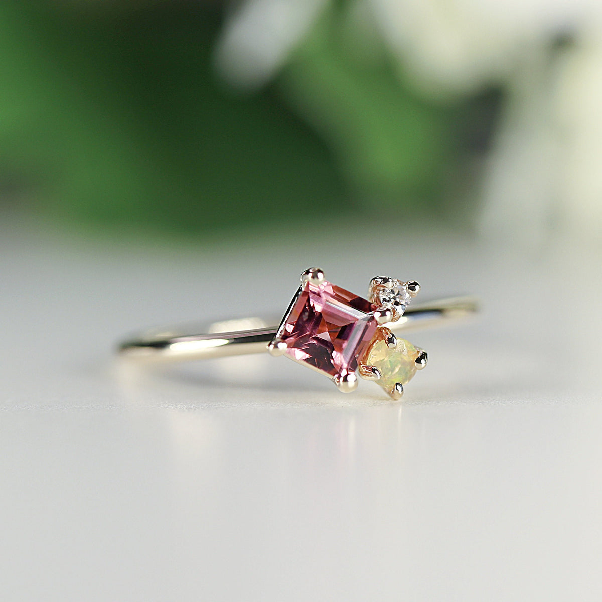 A gold ring with a pink square tourmaline gemstone, a smaller white round gemstone, and a yellow-green square gemstone.