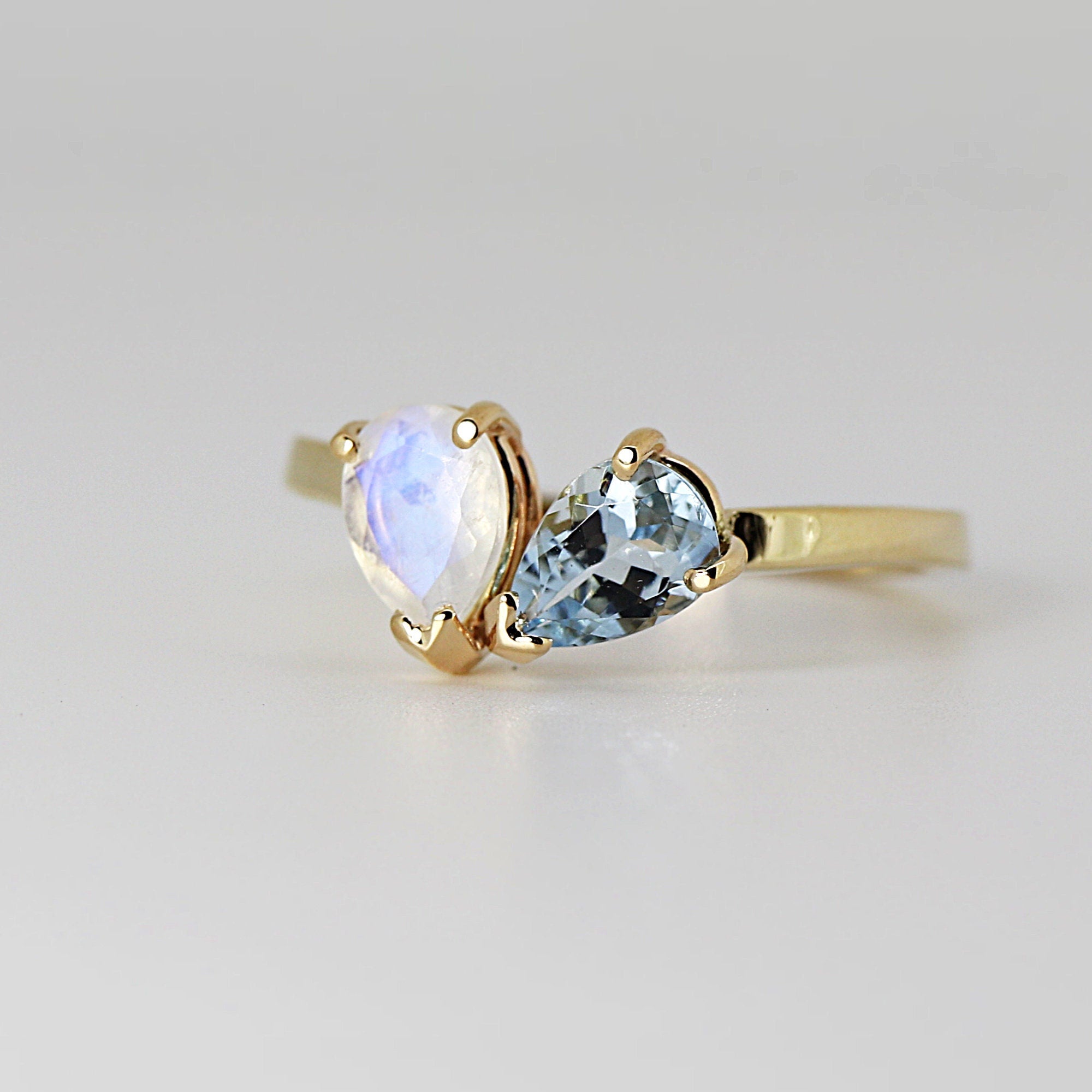 A gold ring featuring two pear-shaped gemstones. One is a luminous, iridescent moonstone, and the other is a pale blue aquamarine. The ring's band and settings are a warm gold color, creating a delicate and elegant design.