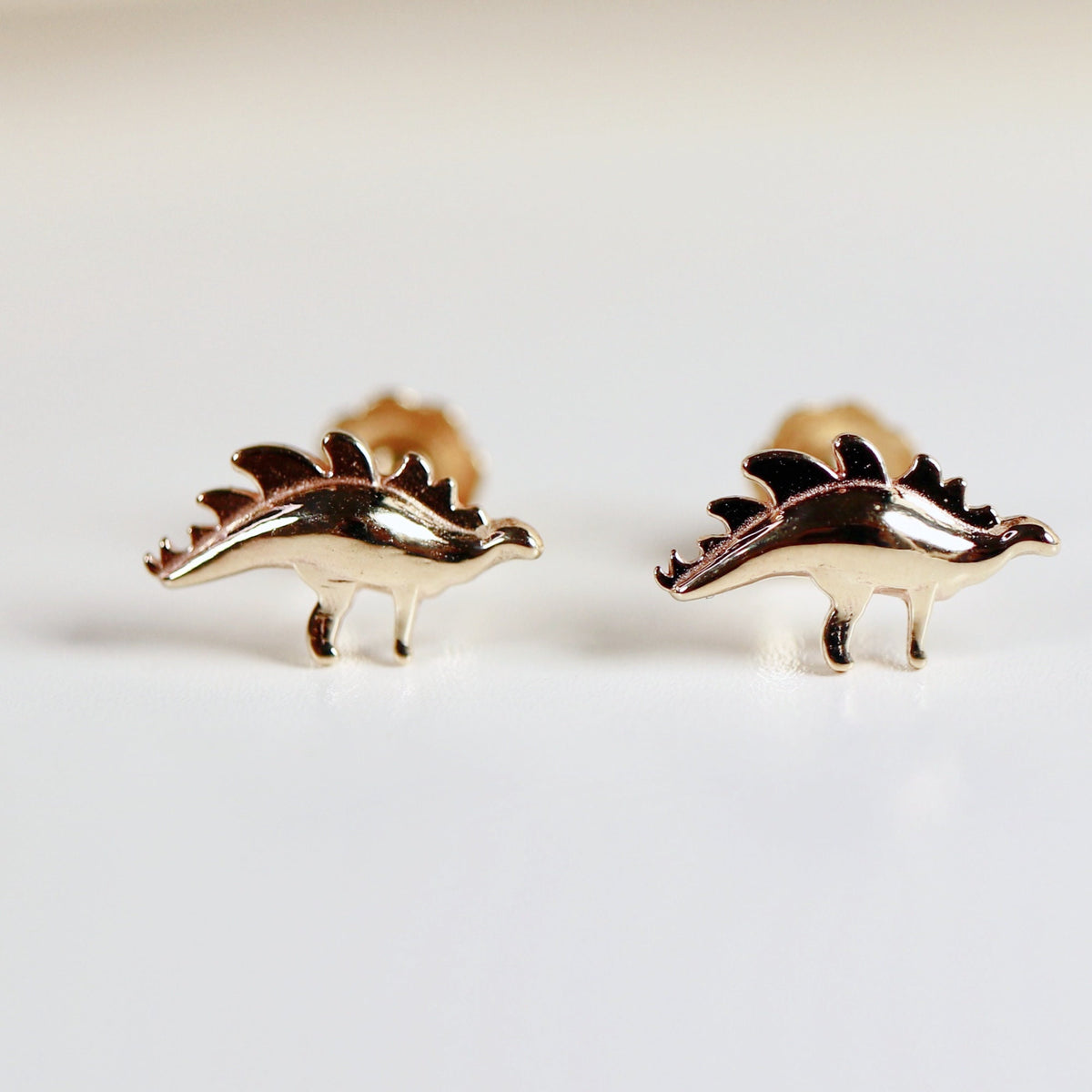 A pair of gold-toned dinosaur stud earrings. The dinosaurs are facing to the right and have a spiky back.