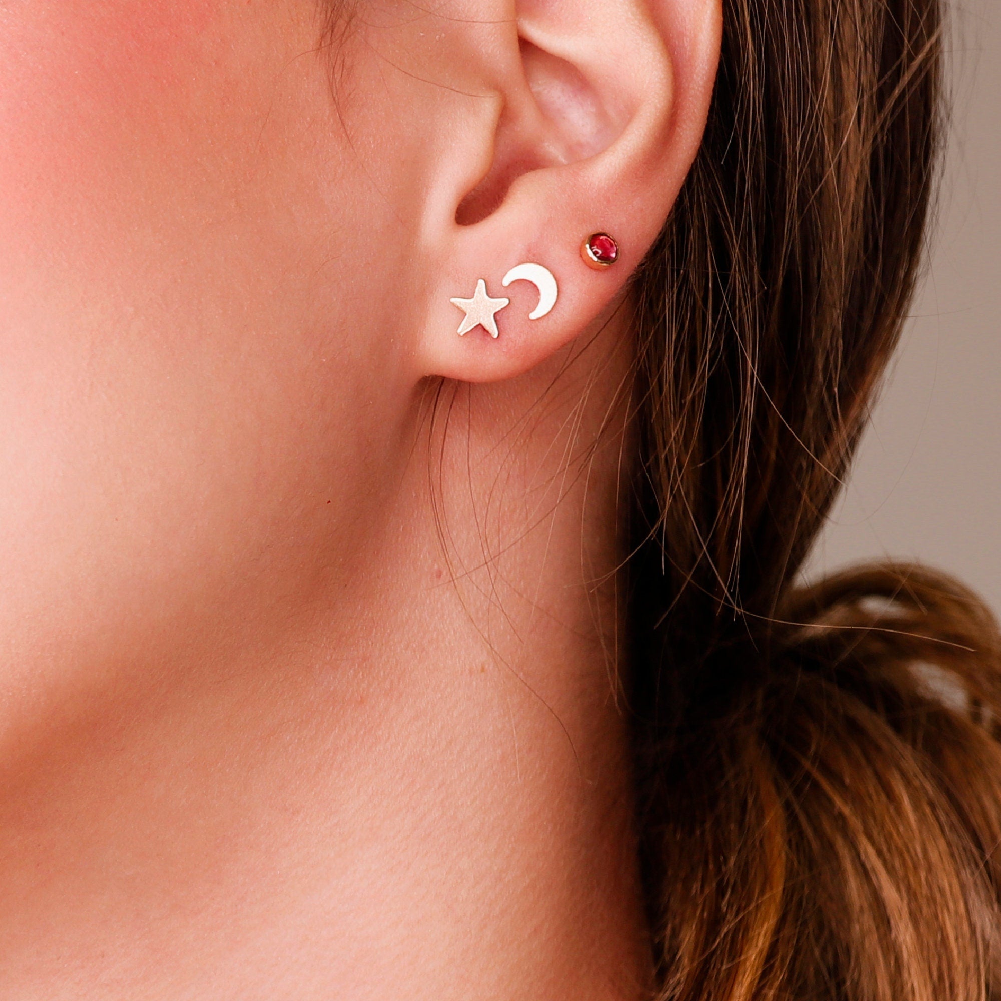 A close-up of a person's ear with three earrings. The earrings are a gold star, a gold crescent moon, and a small red gemstone.Moon and Star Stud Earrings 14k Solid Gold