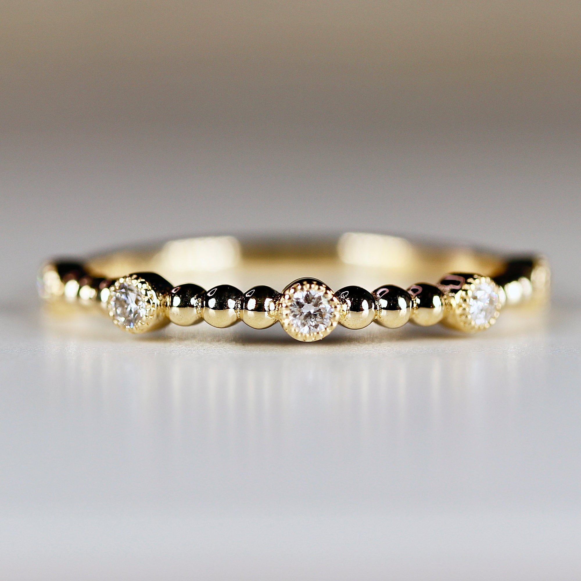 A delicate gold ring featuring a series of small, evenly spaced beads. Three larger, bezel-set diamonds are interspersed among the beads. The ring has a polished, shiny finish, and is set against a soft, neutral background.