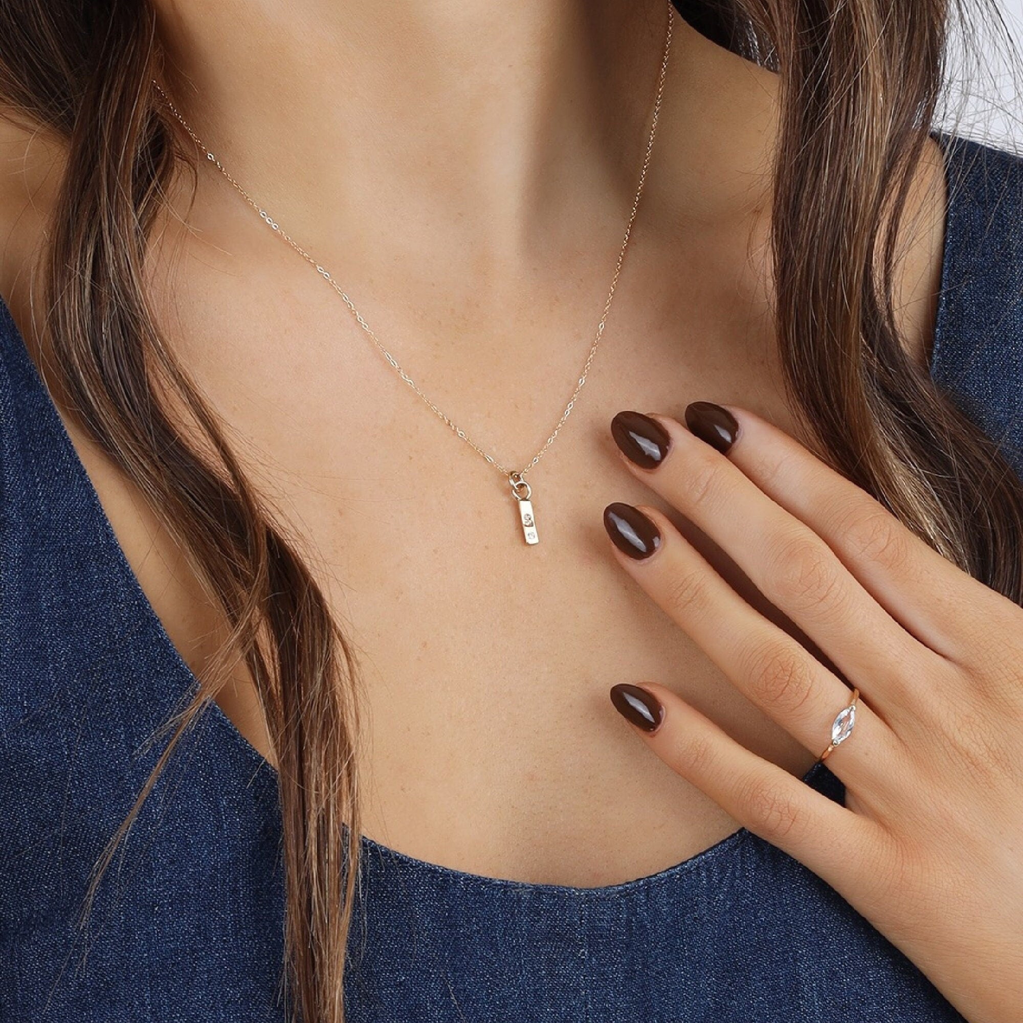 A delicate gold chain necklace with a small, rectangular pendant featuring a single diamond. The necklace is worn by a person with brown hair and dark brown nail polish.