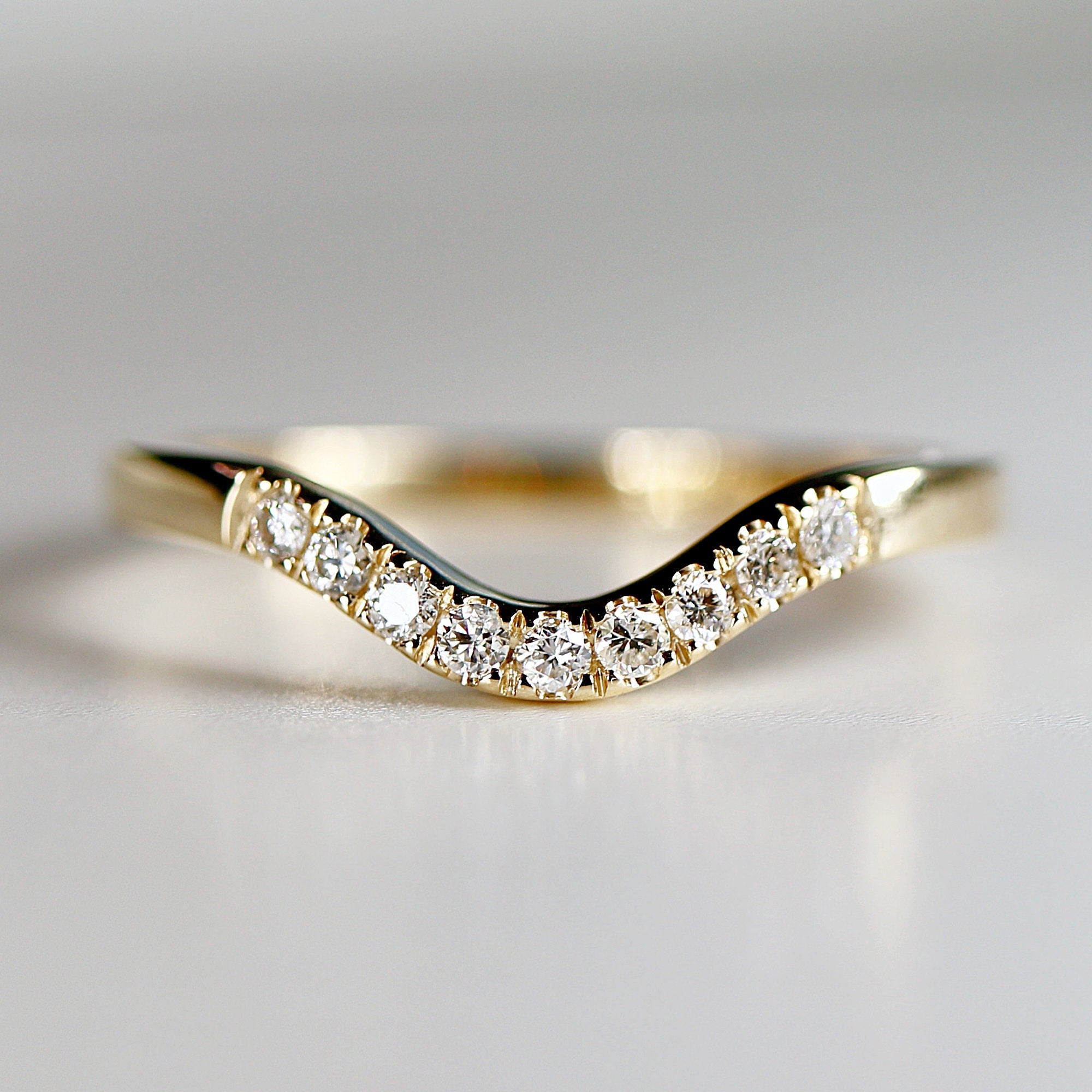 A curved gold ring with a row of small, sparkling diamonds set along the top. The ring has a smooth, polished finish and a delicate, elegant design. The diamonds are clear and catch the light, creating a brilliant effect.