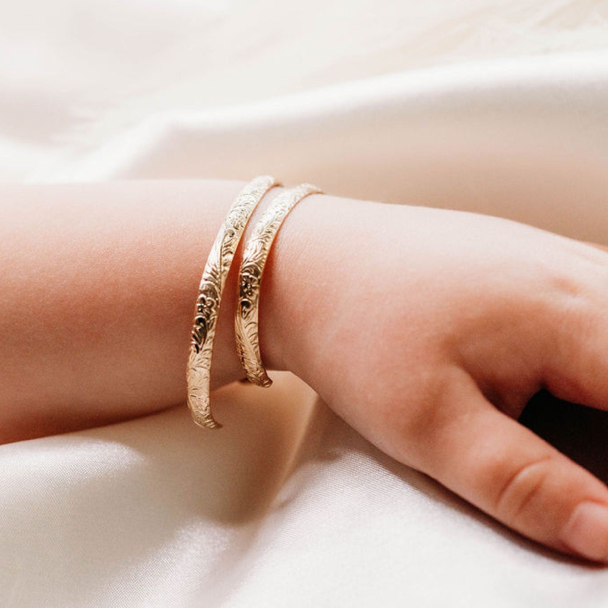 A baby wearing two personalized cuff bracelets with engraved floral and leaf designs, showcasing custom hidden messages inside. Baptism or baby shower jewelry gift.