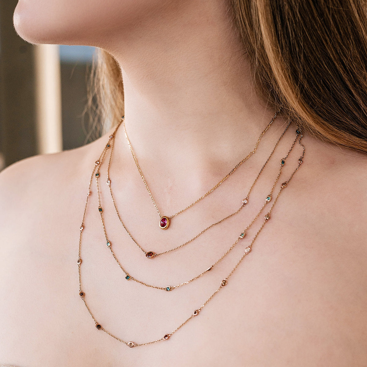 Four delicate gold necklaces of varying lengths. The top necklace features a small, oval-shaped ruby pendant. The other necklaces are adorned with small, colorful gemstones. The necklaces are layered, creating a sophisticated and elegant look.