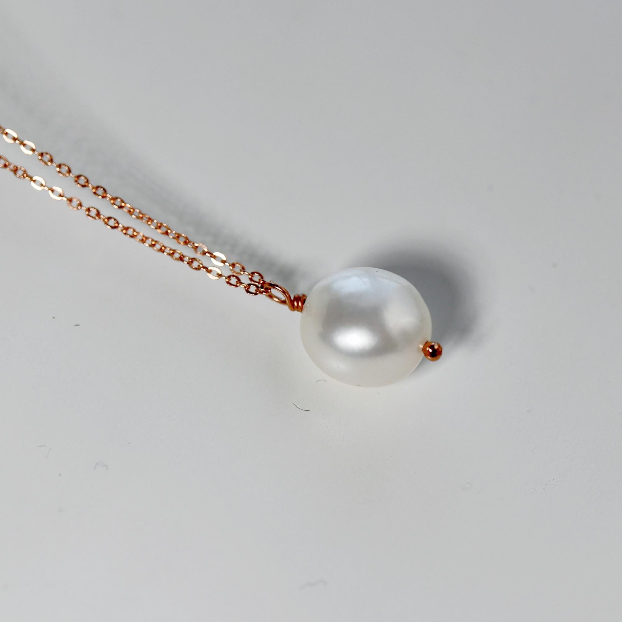 A delicate necklace featuring a large, lustrous white pearl pendant. The pearl is suspended from a fine, rose gold chain. The necklace is displayed against a plain white background, highlighting the pearl's natural beauty.