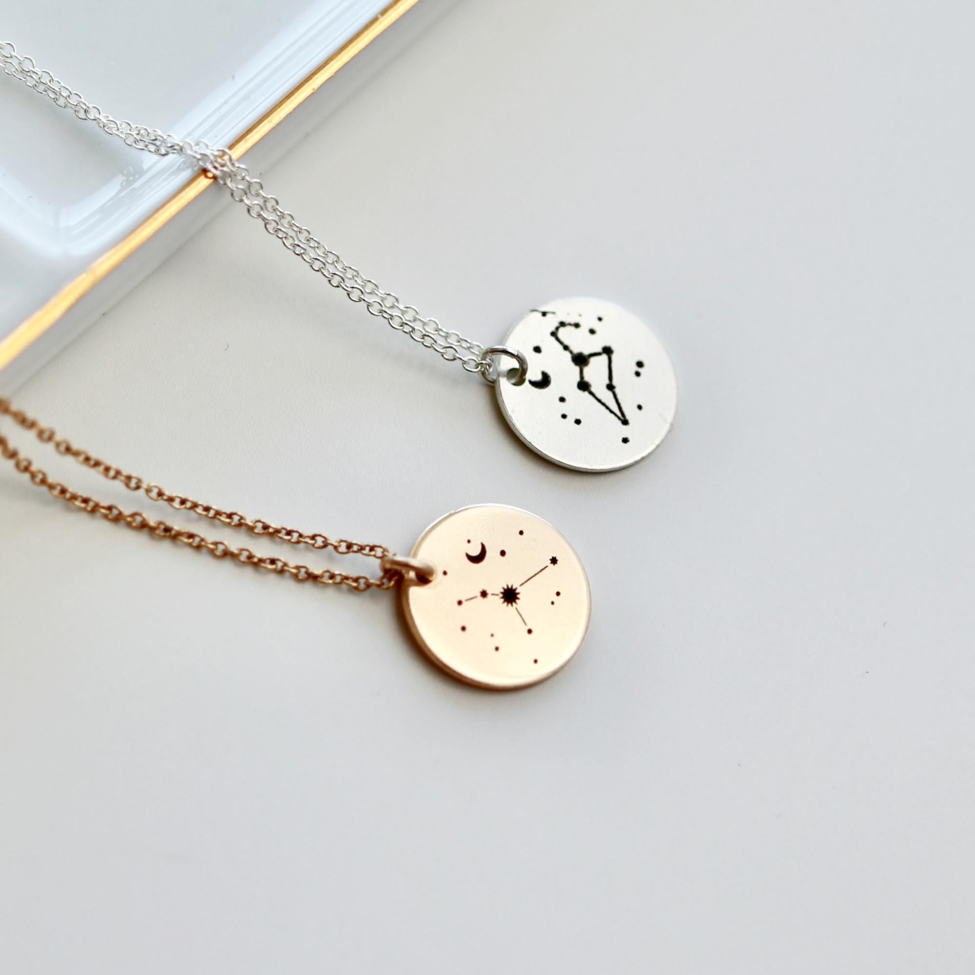 Two delicate necklaces with circular pendants featuring constellation designs. One necklace has a silver chain and a silver pendant with a constellation design. The other necklace has a rose gold chain and a rose gold pendant with a constellation design.