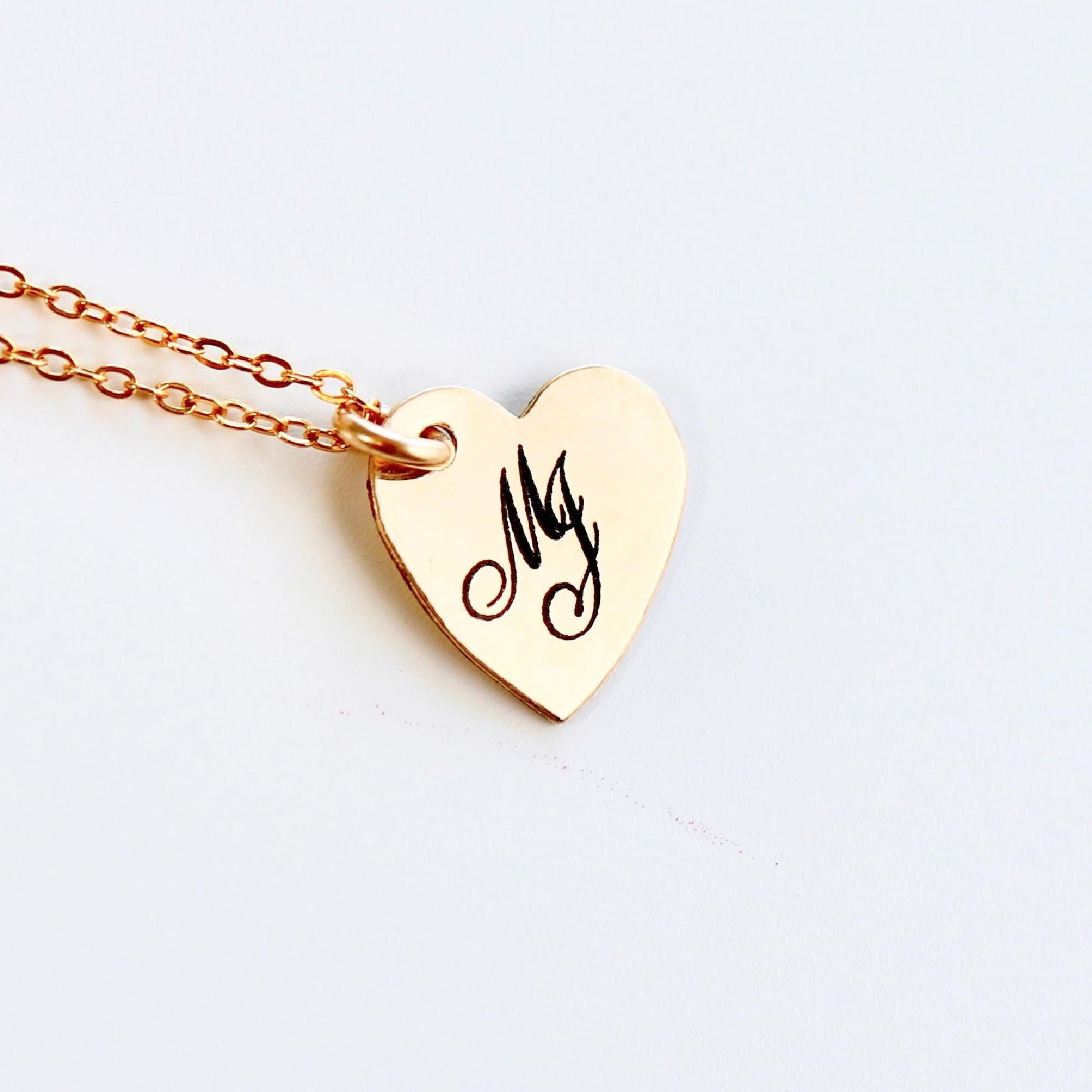 A gold heart-shaped pendant necklace with a delicate chain. The pendant has the letter "M" engraved on it.