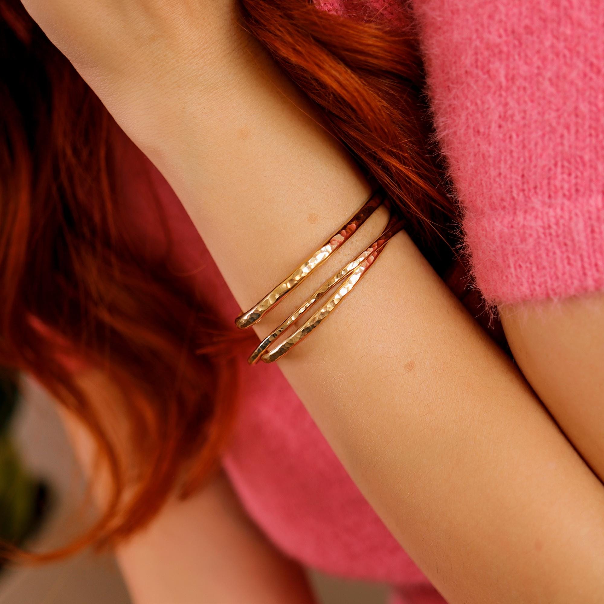 A woman wearing three hammered gold cuff bracelets in varying thicknesses, stacked on her wrist. Gold filled bangles with a personalized engraved message option.
