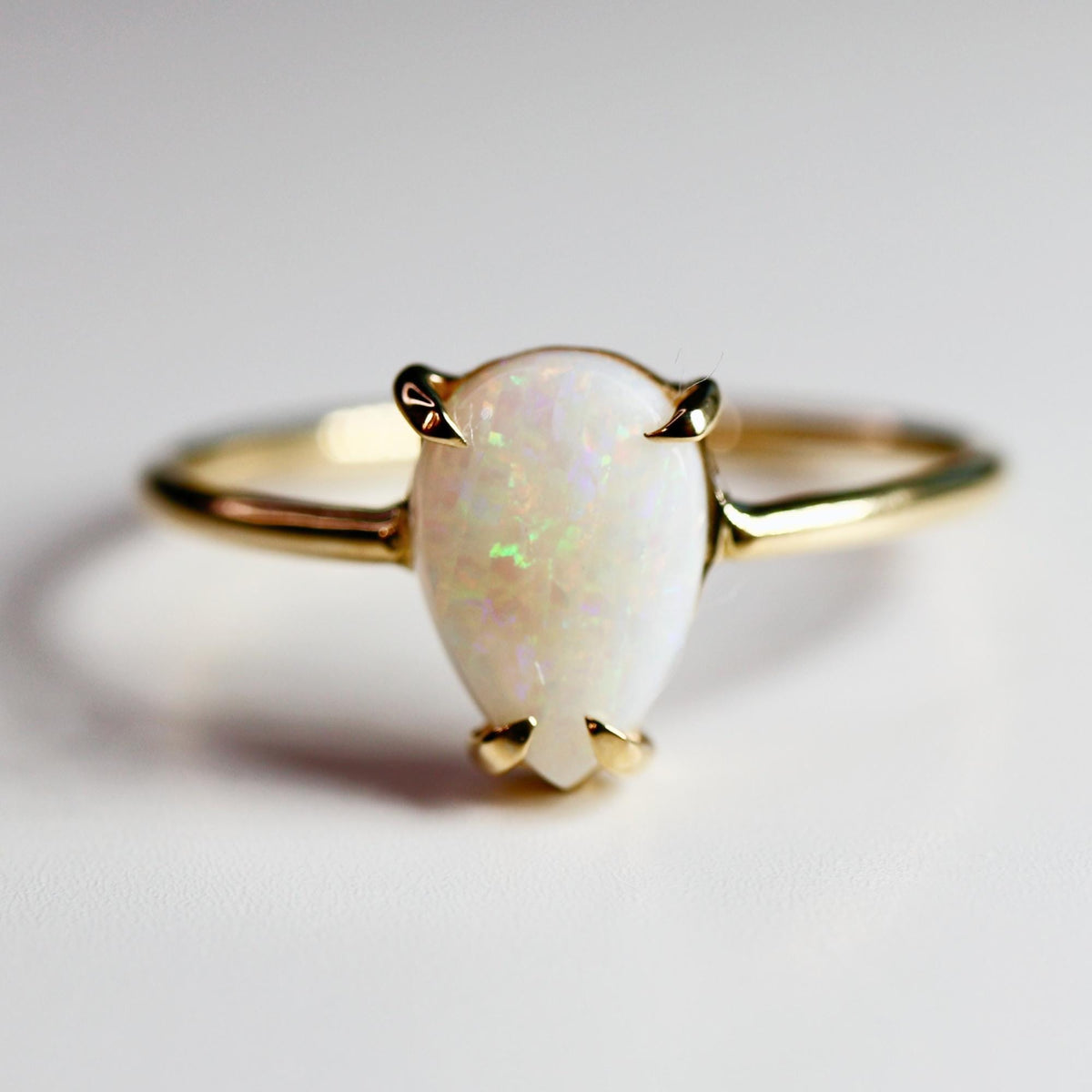 A gold ring featuring a pear-shaped opal gemstone. The opal has a milky white base with iridescent flashes of color. The ring's band and prongs are gold, creating a classic and elegant design. The ring is set against a white background.