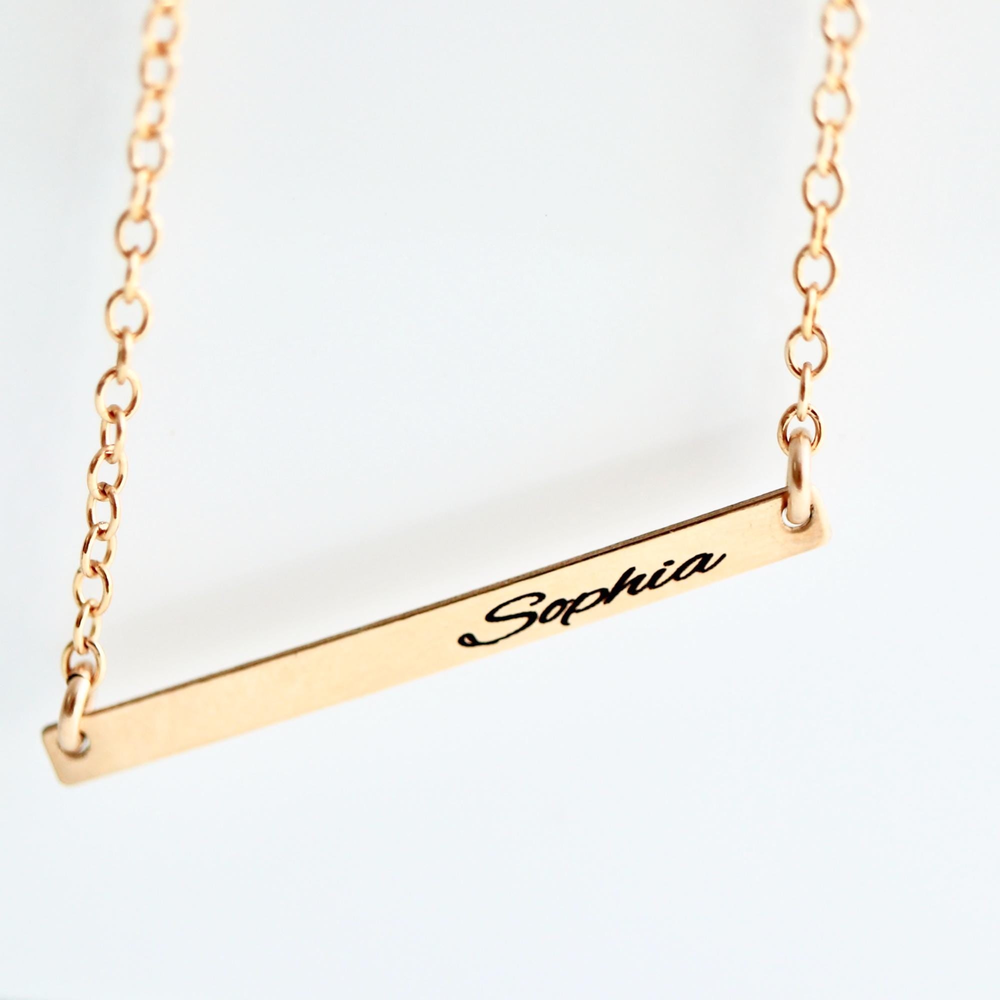 A gold-colored necklace with a delicate chain. The necklace features a horizontal bar pendant with the name "Sophia" engraved in a cursive font. The chain and pendant are a warm, shiny gold.