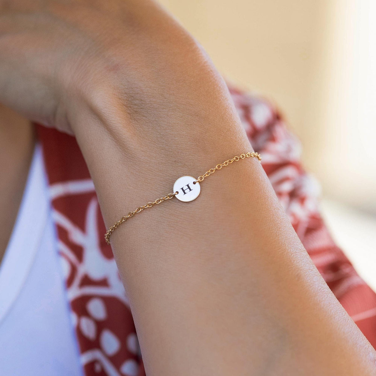 A gold chain bracelet with a small round charm featuring the letter &amp;quot;H&amp;quot; in black.