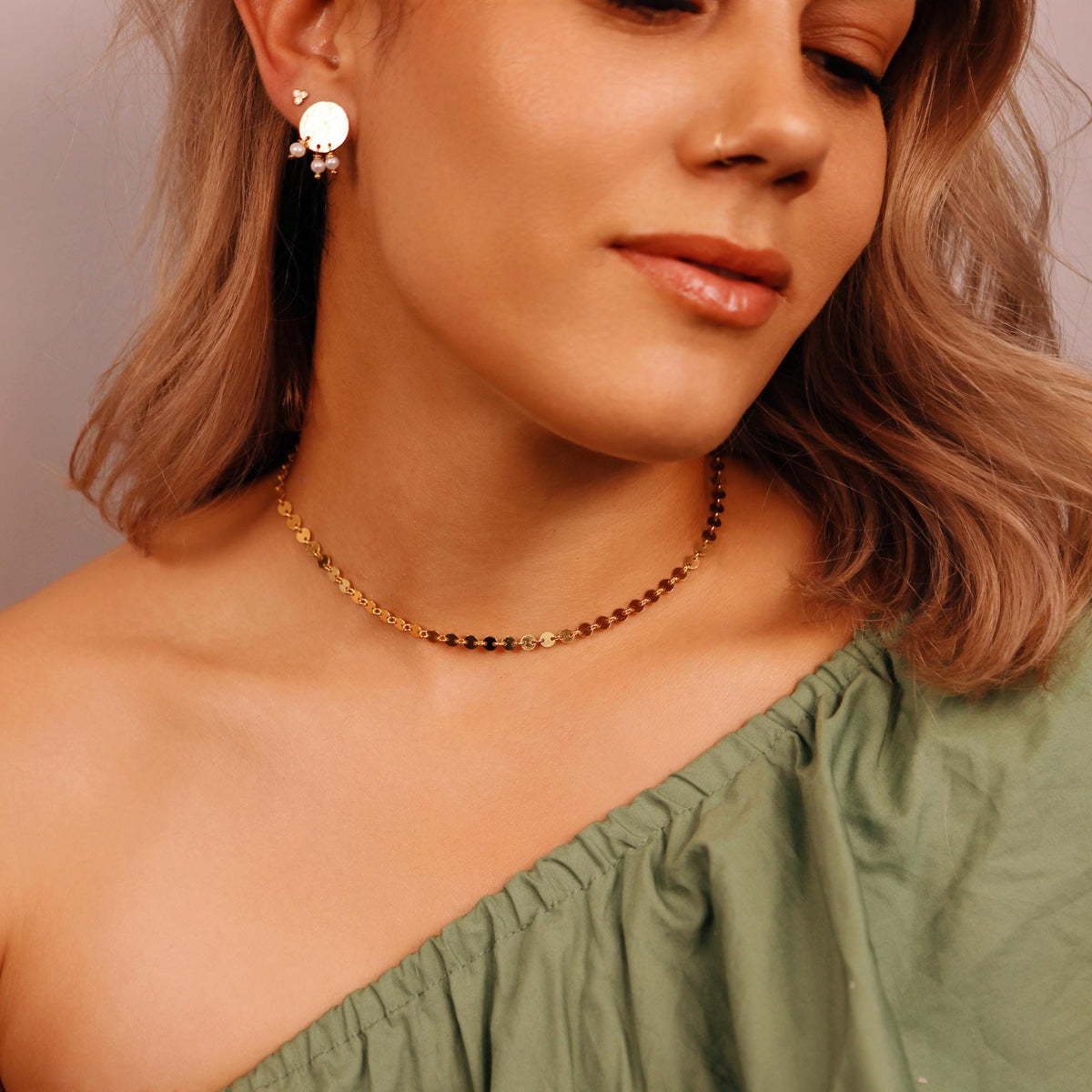 A gold chain choker necklace with small round links. The necklace is worn by a person with long blonde hair. The person is wearing a green shirt.